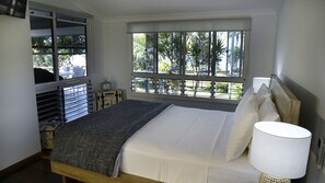 A bedroom with a bed, a window with a view of palm trees, a bedside table with a lamp, and a storage unit with suitcases.