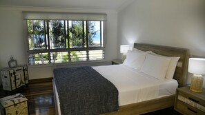 A bedroom with a bed, a window with a view of palm trees, a bedside table with a lamp, and a nightstand with a lamp. - Ohana - Beachfront Holiday House (South Mission Beach)
