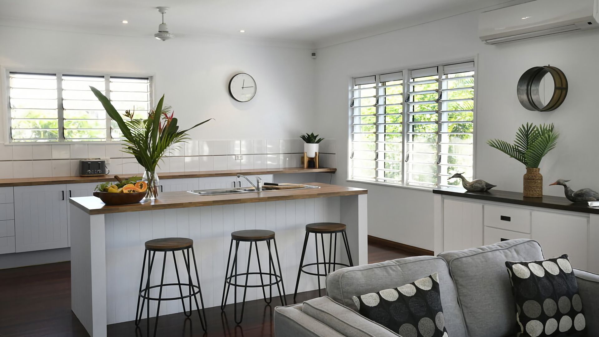 A modern kitchen with a central island, bar stools, a dining area with a sofa, and large windows with blinds.