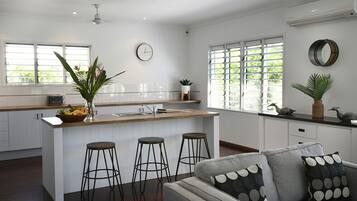 A modern kitchen with a central island, bar stools, a dining area with a sofa, and large windows with blinds.