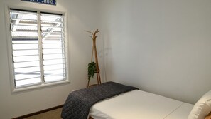 Une chambre à coucher avec un lit, une fenêtre avec des stores et un support à plantes en bois.