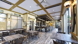 A restaurant interior with wooden tables and chairs, a bar area, and large windows allowing natural light.