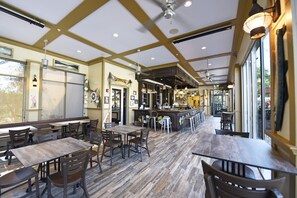 A restaurant interior with wooden tables and chairs, a bar area, and large windows allowing natural light.
