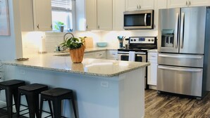 A modern kitchen with white cabinets, stainless steel appliances, a large island with stools, and a wooden floor. - Yellow Door Cottage In The Heart of Bay St. Louis (Bay St. Louis)