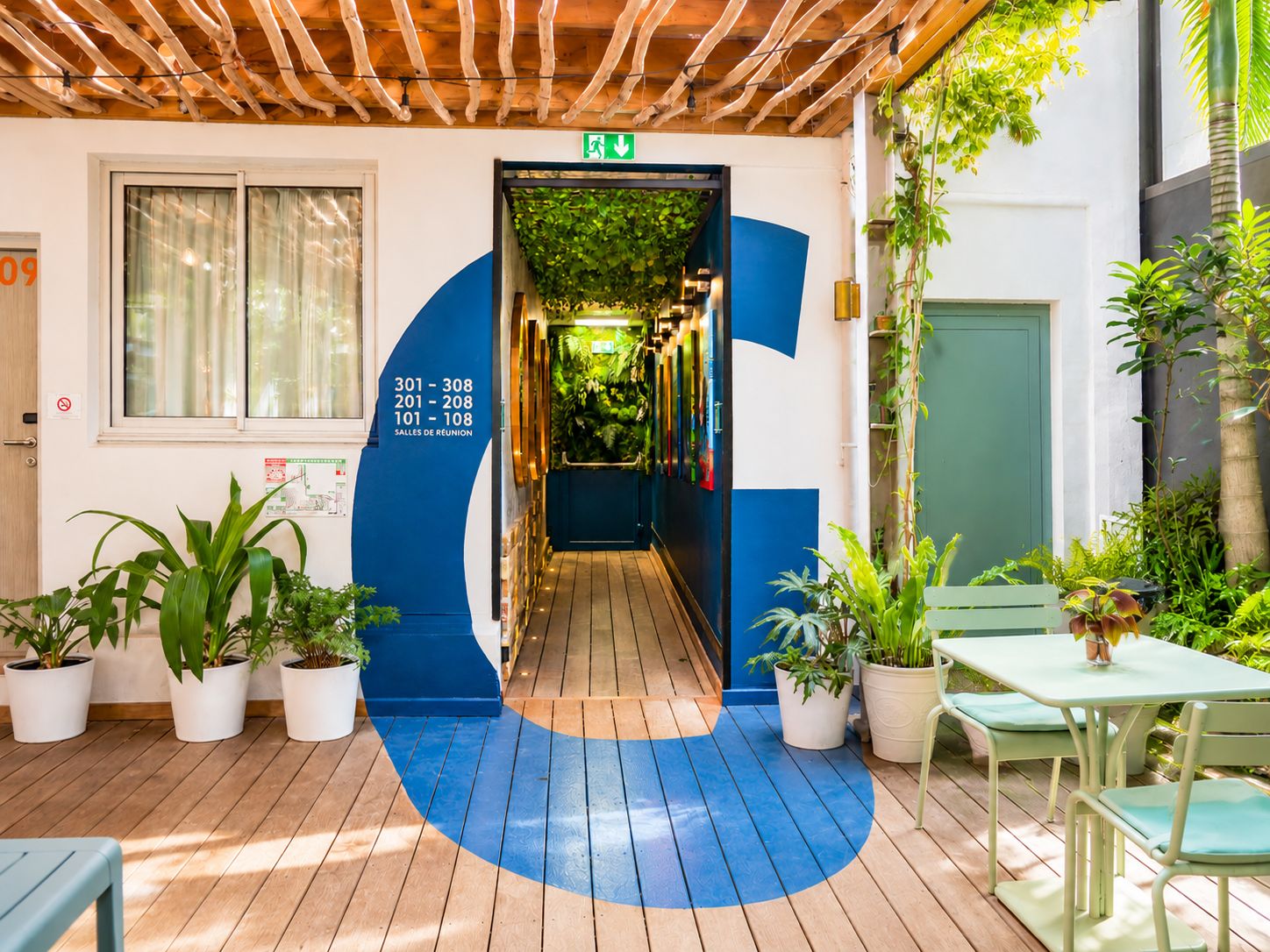 A wooden deck with potted plants, a blue painted wall with room numbers, and a green door.