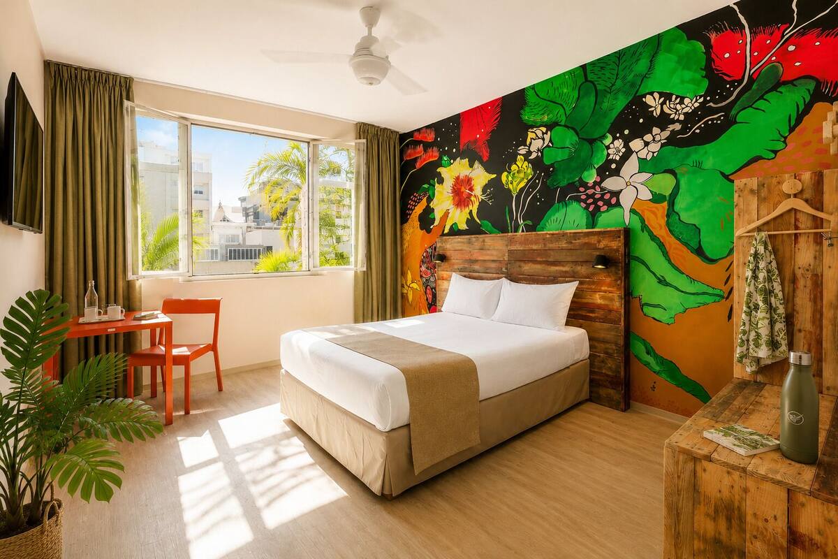 Ein Hotelzimmer mit Bett, Schreibtisch, Stuhl, einem Fenster mit Vorhängen, einem Deckenventilator und einem farbenfrohen Wandgemälde.
