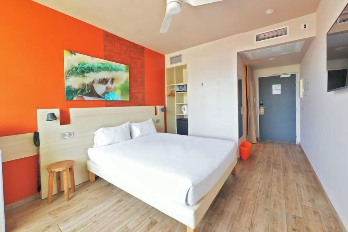 A hotel room with a bed, a small stool, a painting on the wall, and a door leading to another room.