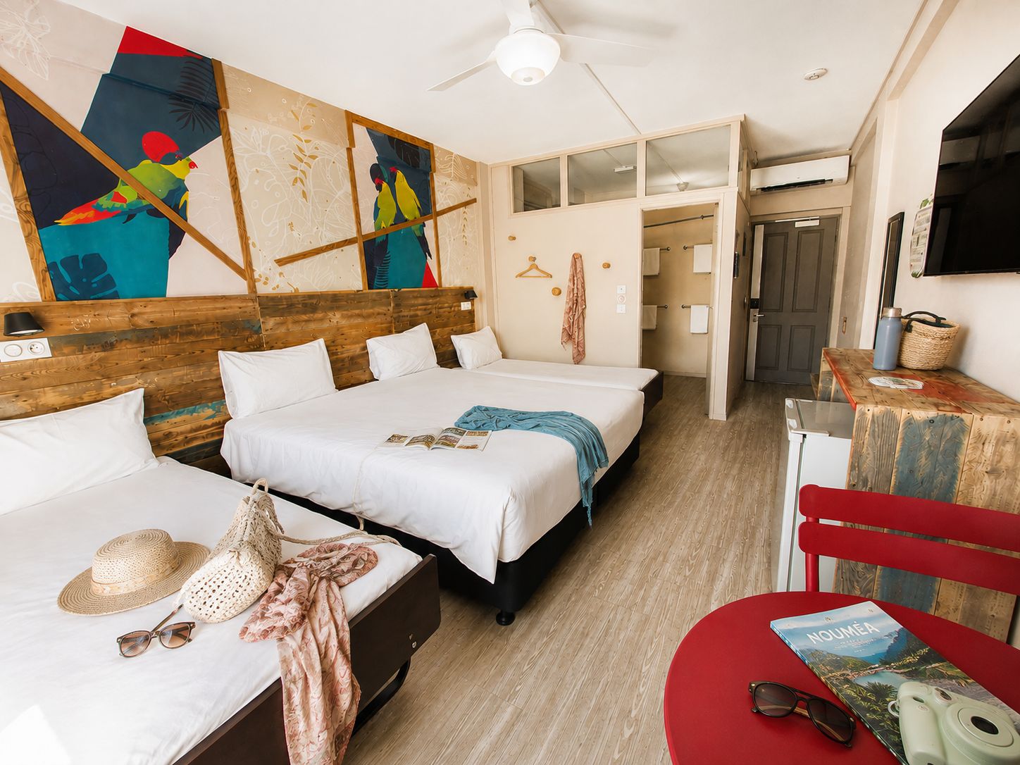 A hotel room with two beds, a wooden headboard, a ceiling fan, a wall with colorful artwork, and a red chair with a book.