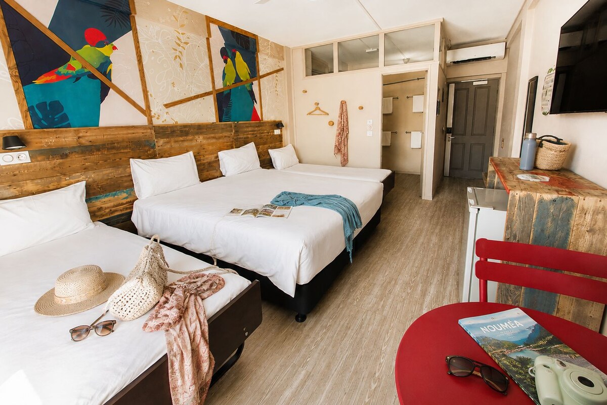 Ein Hotelzimmer mit zwei Betten, einem Holzkopfteil, einem Deckenventilator, einer Wand mit farbenfroher Kunst und einem roten Sessel mit einem Buch.