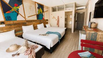 A hotel room with two beds, a wooden headboard, a ceiling fan, a wall with colorful artwork, and a red chair with a book.