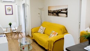 Un divano giallo con un cuscino, un tavolino in vetro, sedie bianche e un quadro appeso al muro.