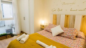 Un letto rifatto con lenzuola a motivi floreali e una coperta color senape, una lampada da comodino e una finestra con tende.