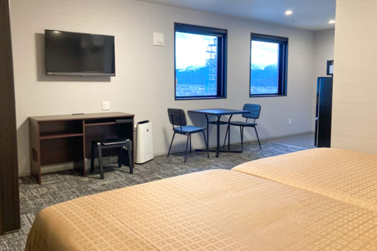 A hotel room with a bed, a desk, two chairs, a television, and windows with a city view.