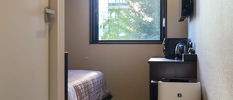 Una habitación de hotel pequeña con una cama, un escritorio con cafetera, un refrigerador y una ventana con vista a edificios y árboles.