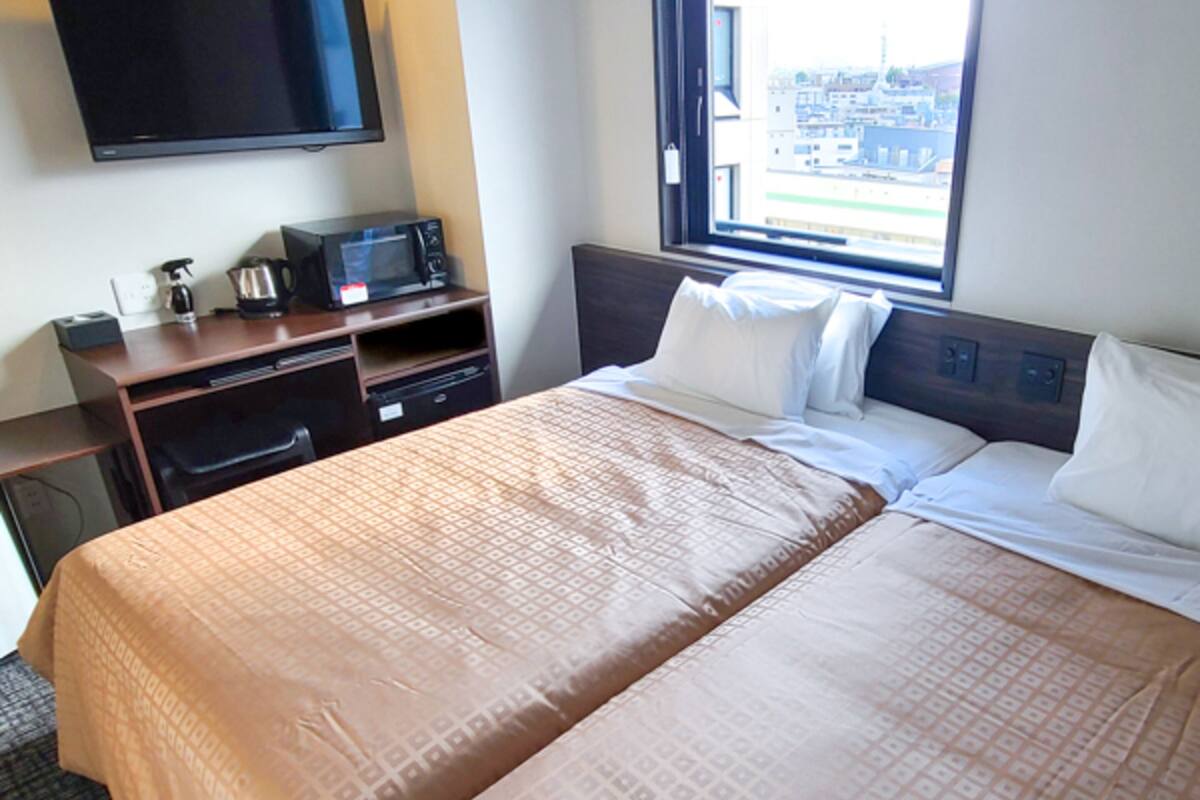 A hotel room with a large bed, a TV mounted on the wall, a microwave on a wooden table, and a window with a city view.
