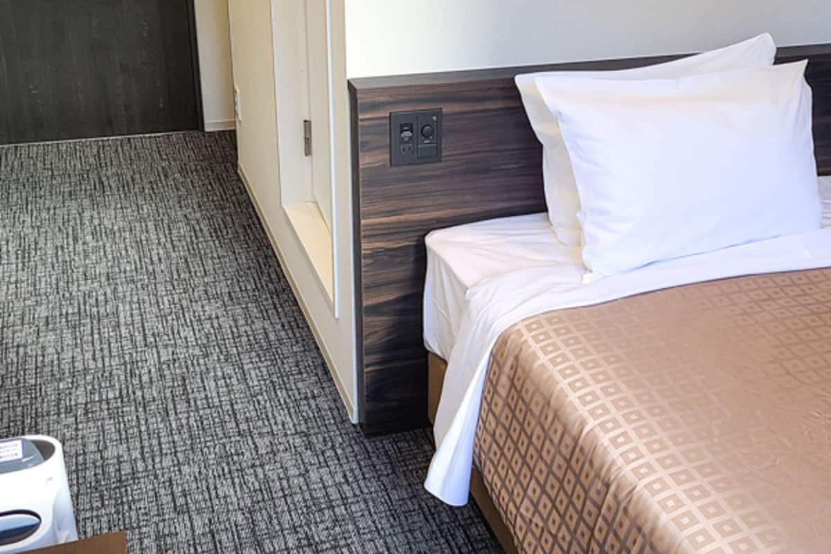 A hotel room with a bed, a carpeted floor, a wooden headboard, and a white wall.