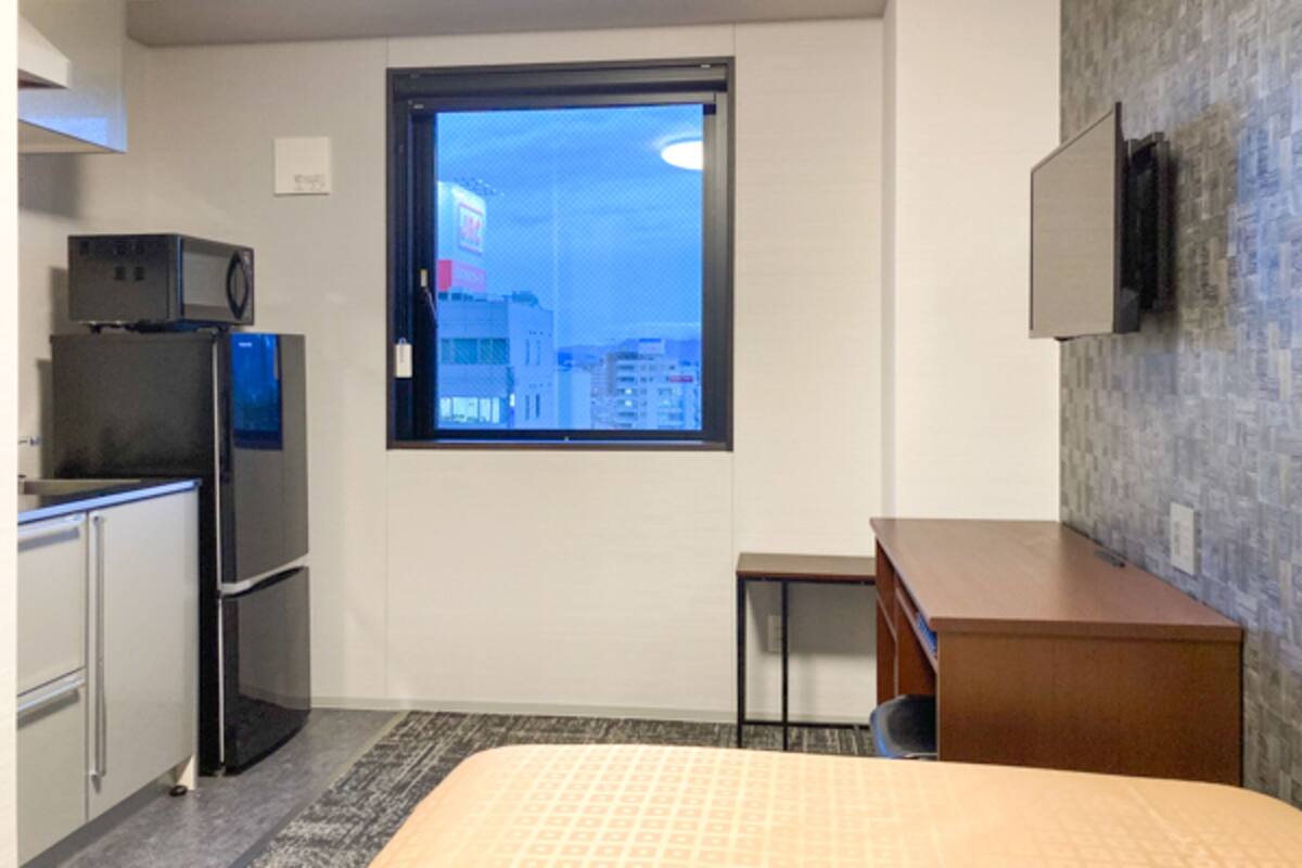 A compact hotel room with a bed, a small kitchenette, a window with a city view, and a flat-screen TV mounted on the wall.