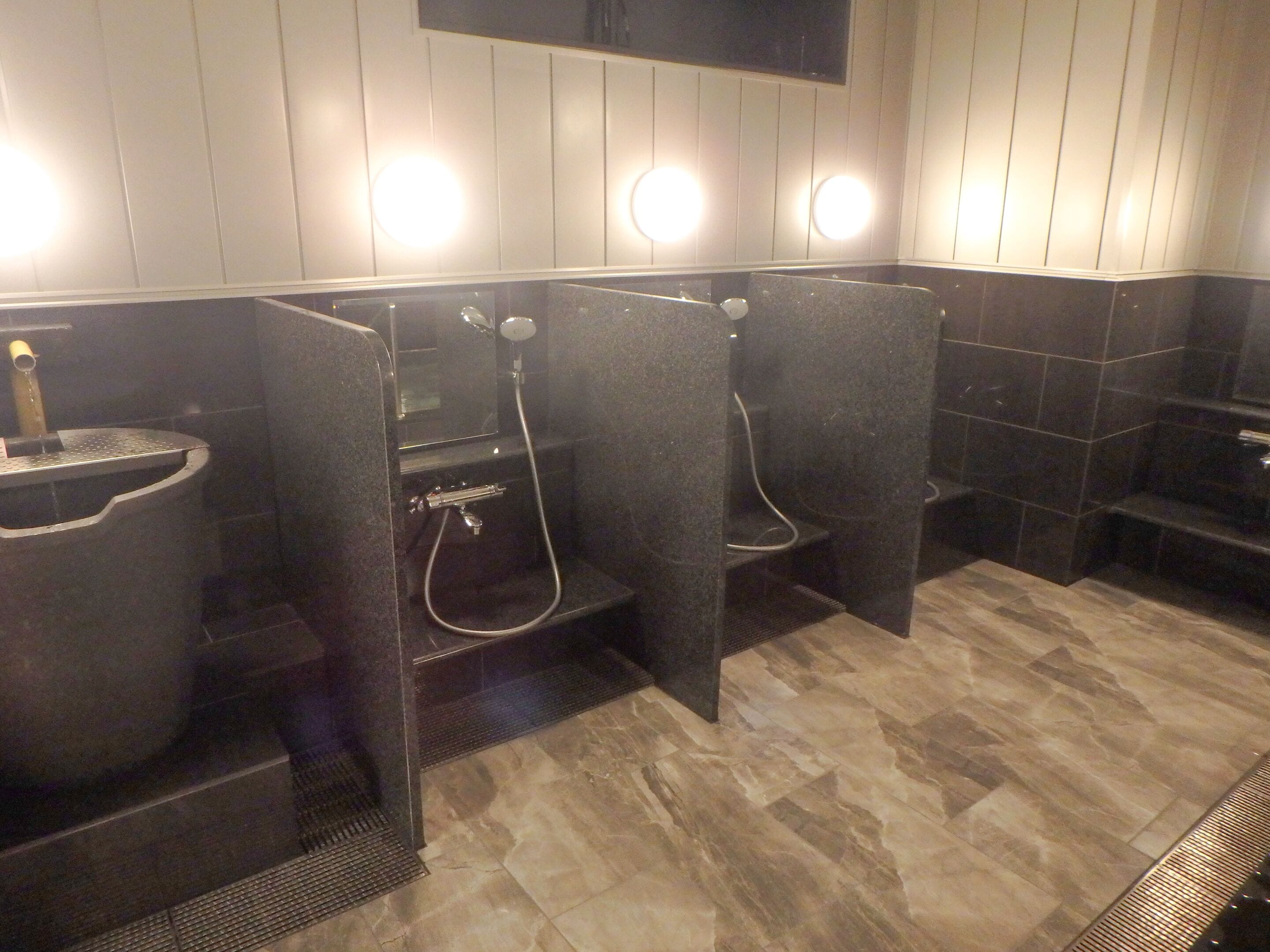 A public restroom with three stalls, each equipped with a toilet, a sink, and a shower. The floor is tiled, and there are two illuminated wall sconces above the stalls.