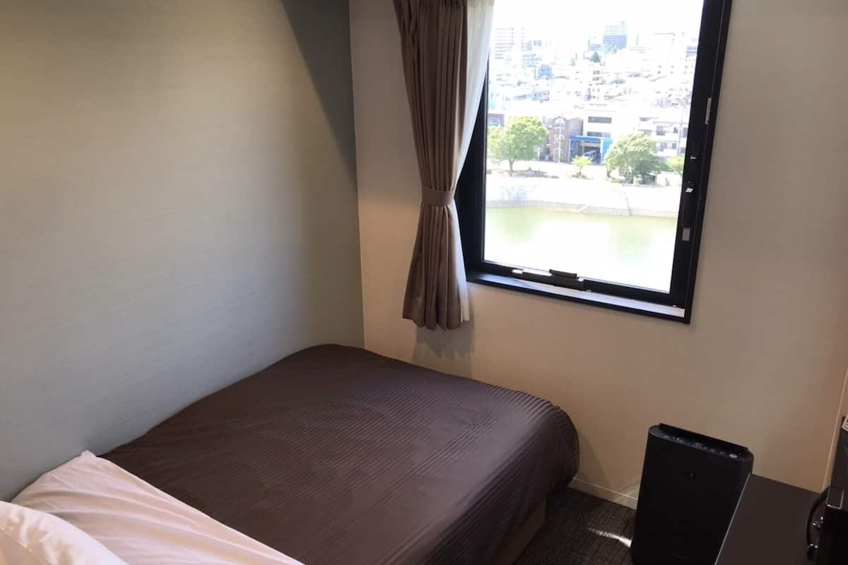 Een hotelkamer met een enkel bed, een raam met gordijnen, een bureau met een zwarte speaker en uitzicht op gebouwen.