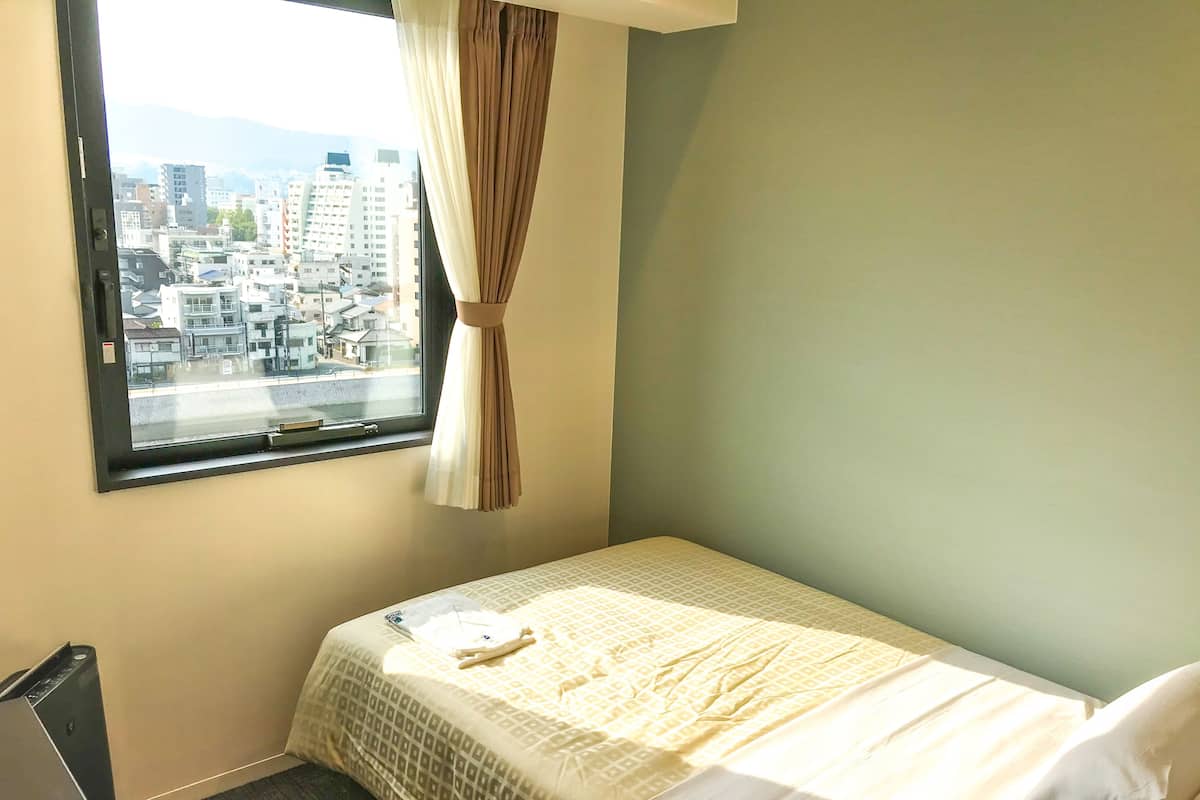 Een kleine hotelkamer met een enkel bed, een raam met gordijn en uitzicht op een stadsbeeld.