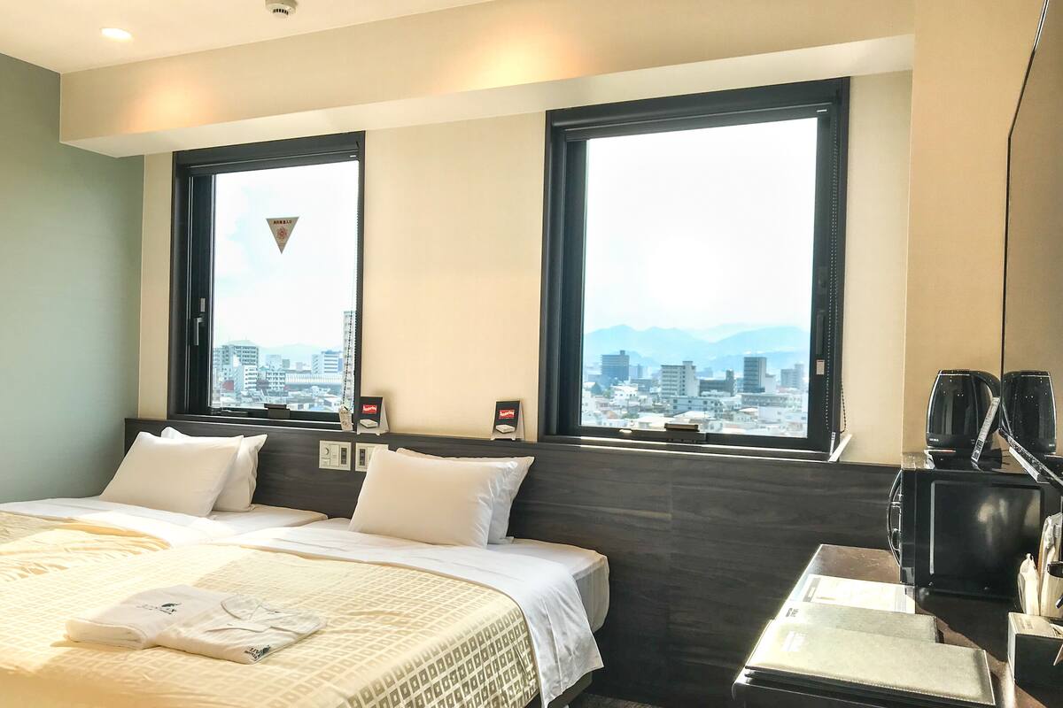 Een hotelkamer met een bed, twee ramen met uitzicht op de stad, een bureau met een koffiezetapparaat, en een spiegel.