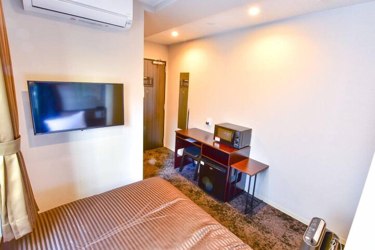 A hotel room with a bed, a flat-screen TV, a desk with a computer, a microwave, and an air conditioning unit.