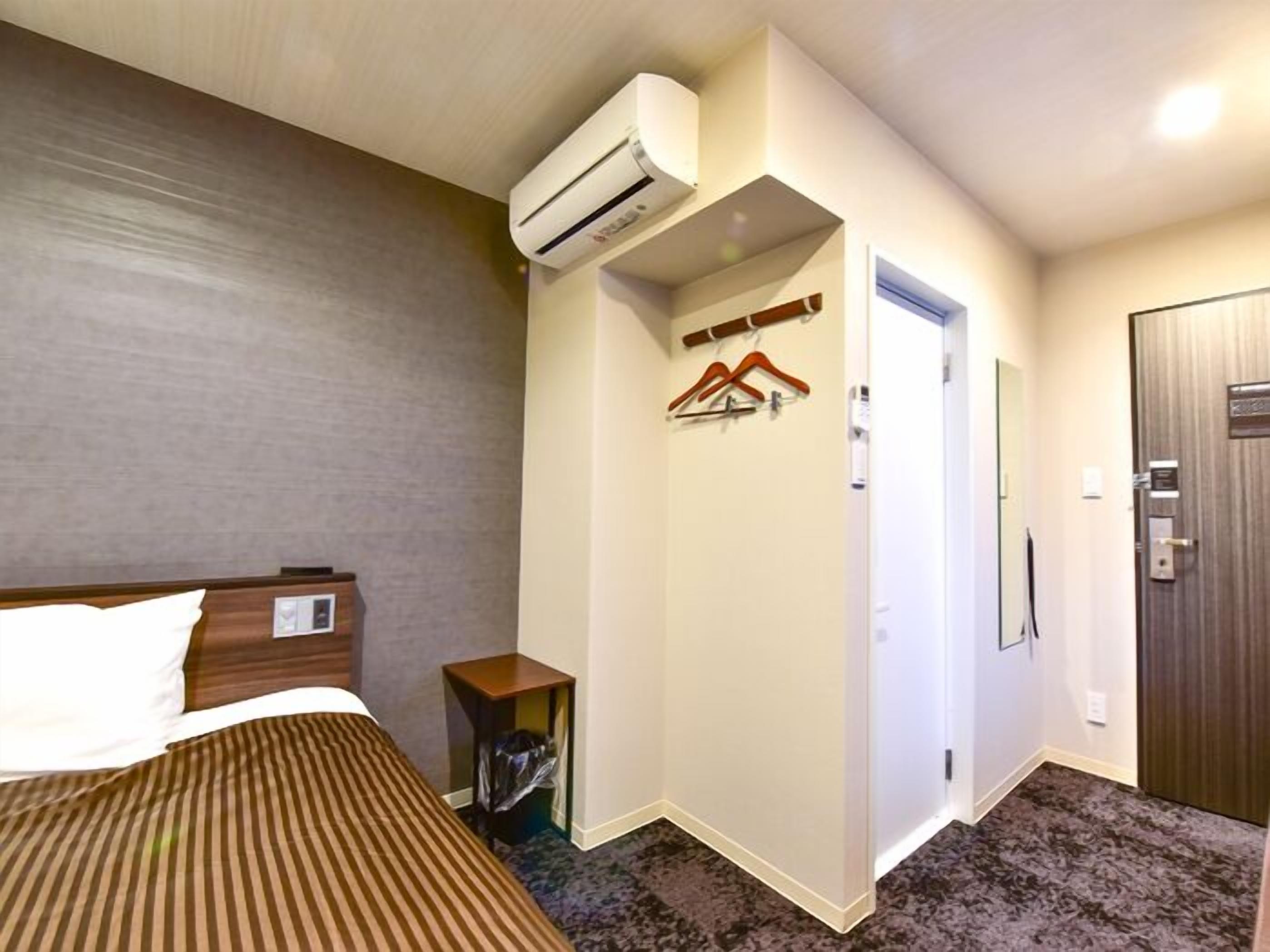 Una habitación de hotel con cama, unidad de aire acondicionado, perchero, mesita y una puerta.