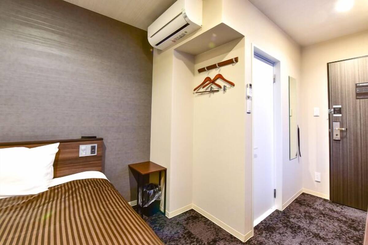 A hotel room with a bed, an air conditioning unit, a coat rack, a small table, and a door.