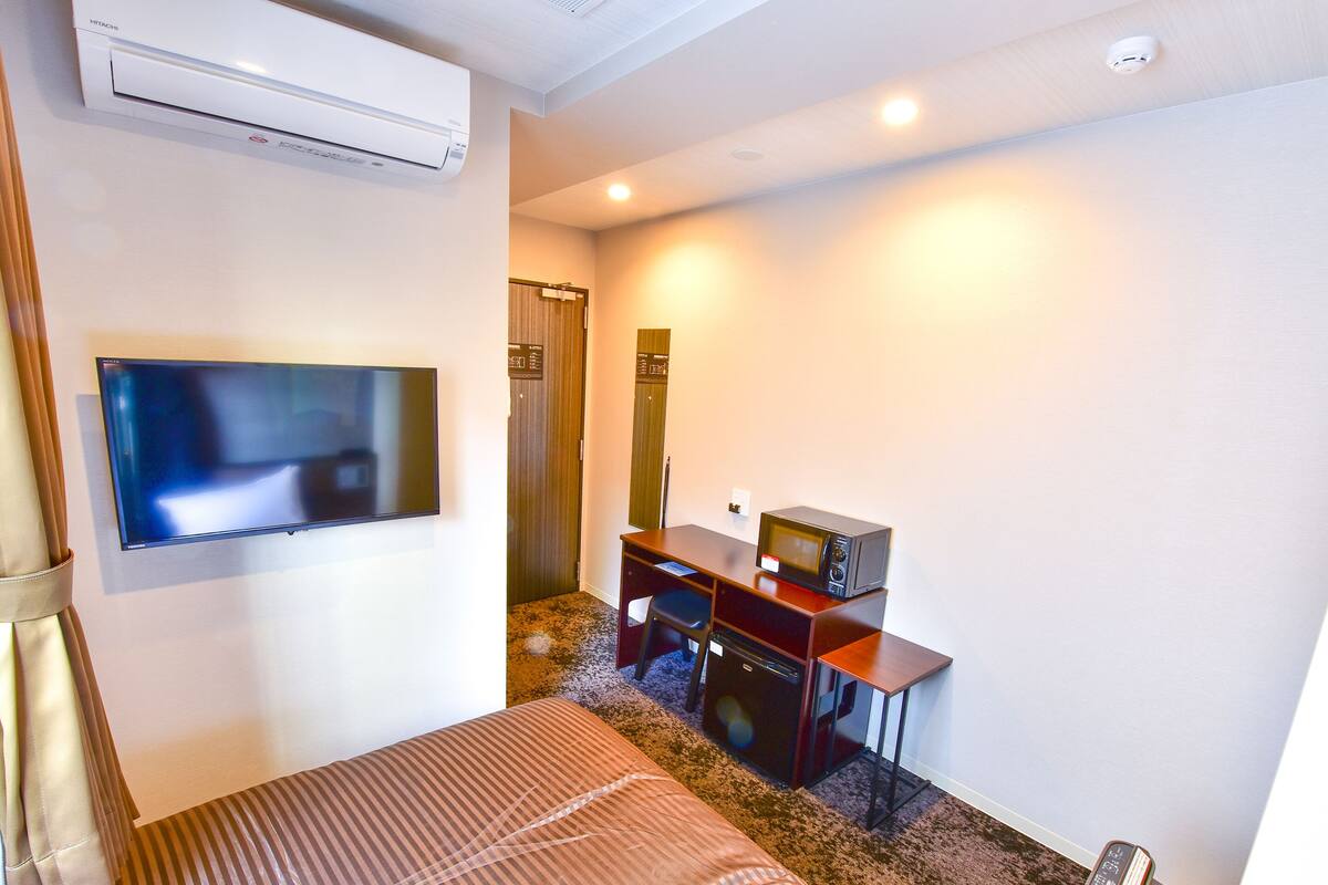 A hotel room with a bed, a desk with a microwave, a television, and an air conditioning unit.