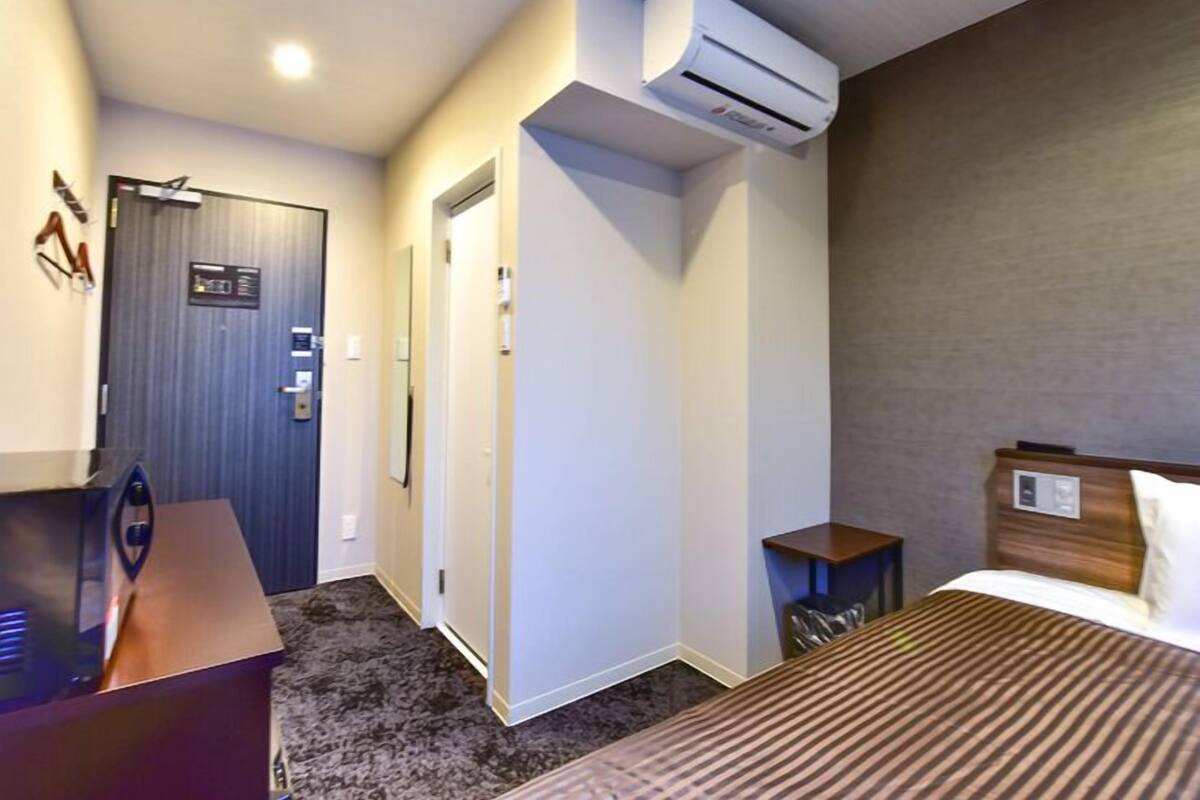 A hotel room with a bed, a desk, an air conditioning unit, a door, and a small table.
