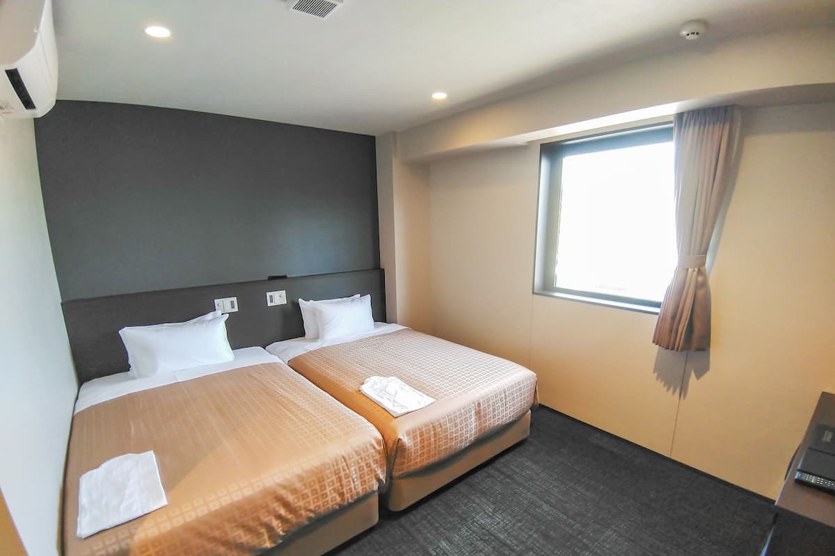 Een hotelkamer met twee bedden, een raam met gordijnen, een donker hoofdeinde en een klein bureau met een computer.