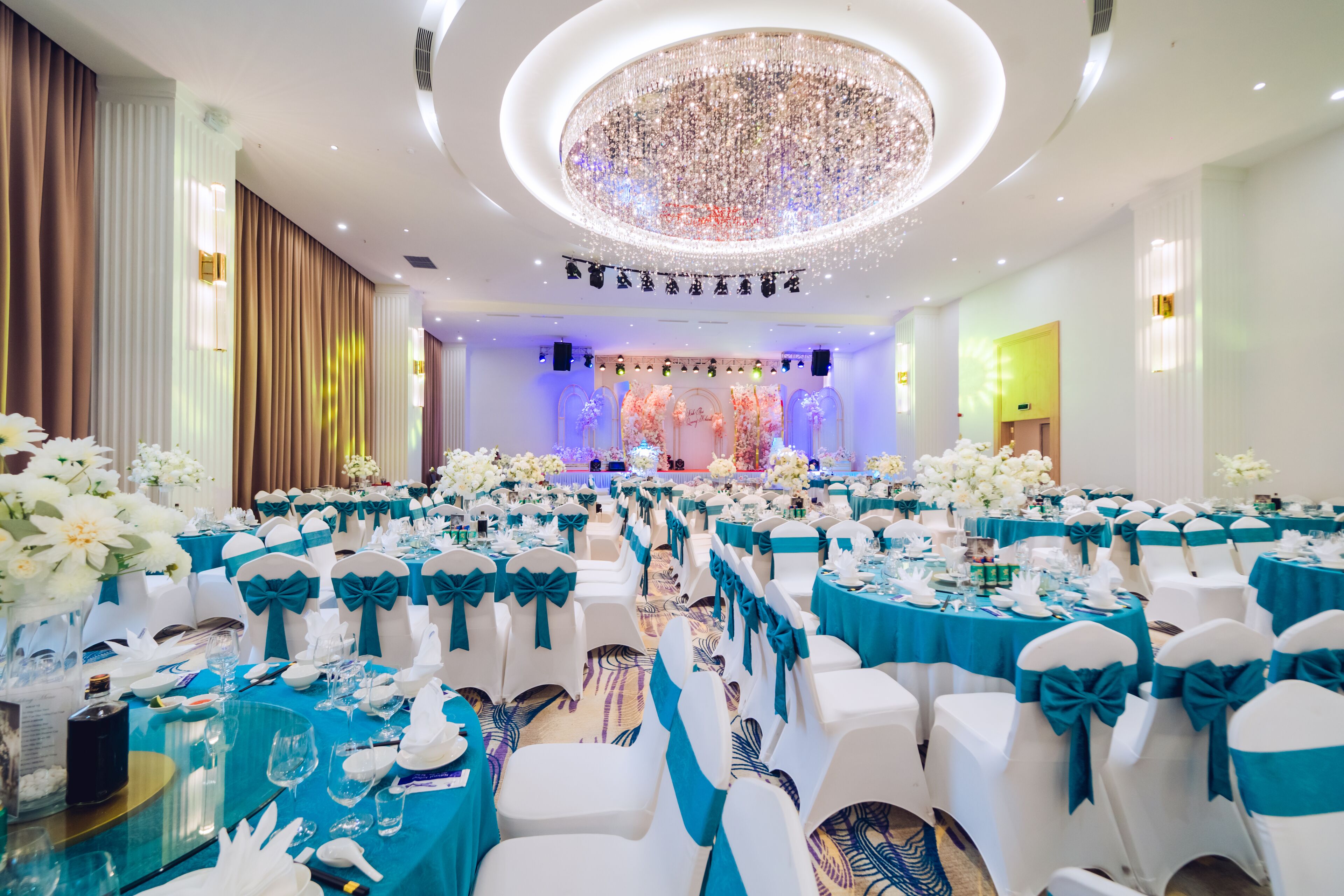 A banquet hall with rows of tables set for a formal event, featuring white chairs with teal sashes and a large, ornate chandelier.