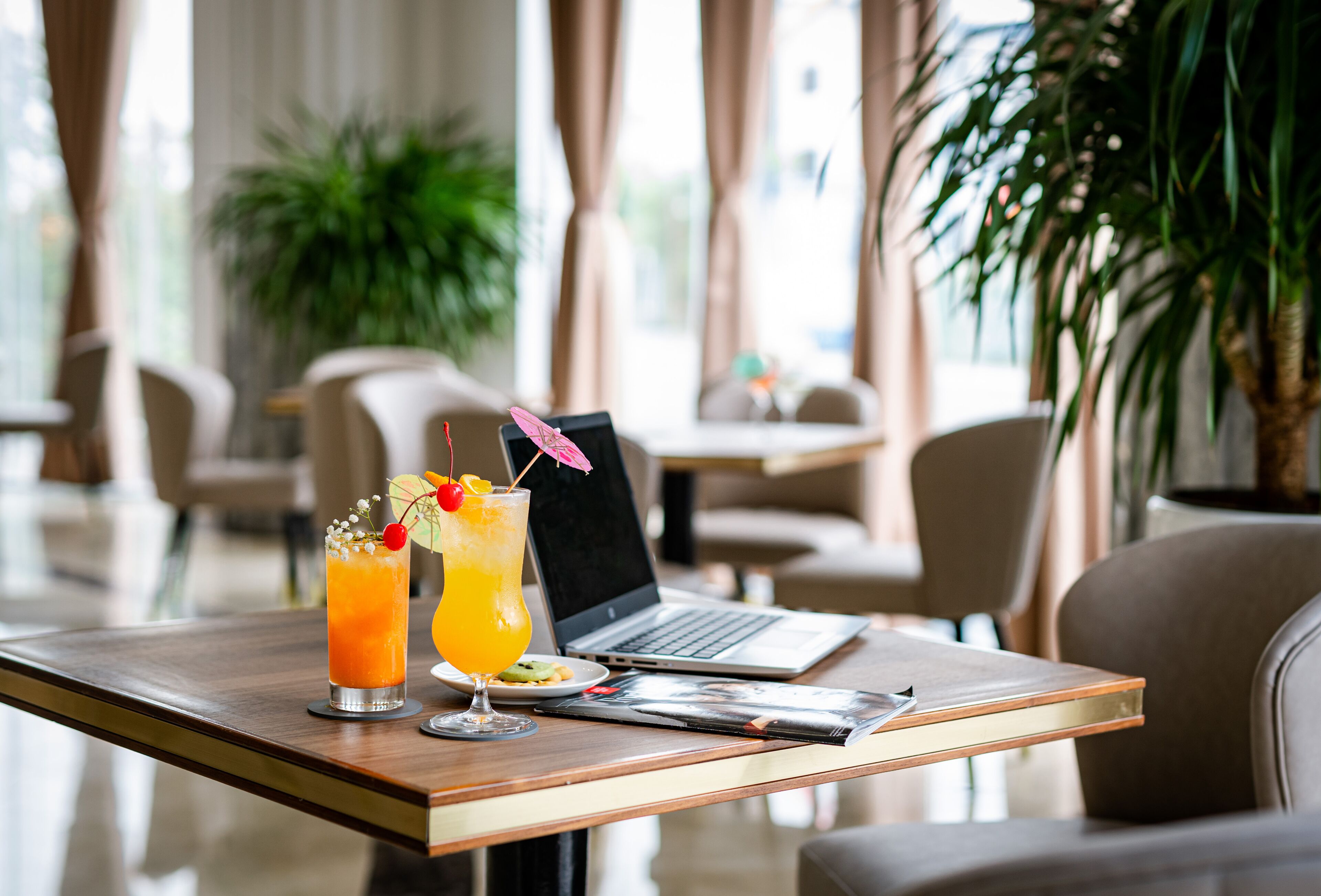 A laptop on a tray table in a hotel room with two drinks and a magazine.