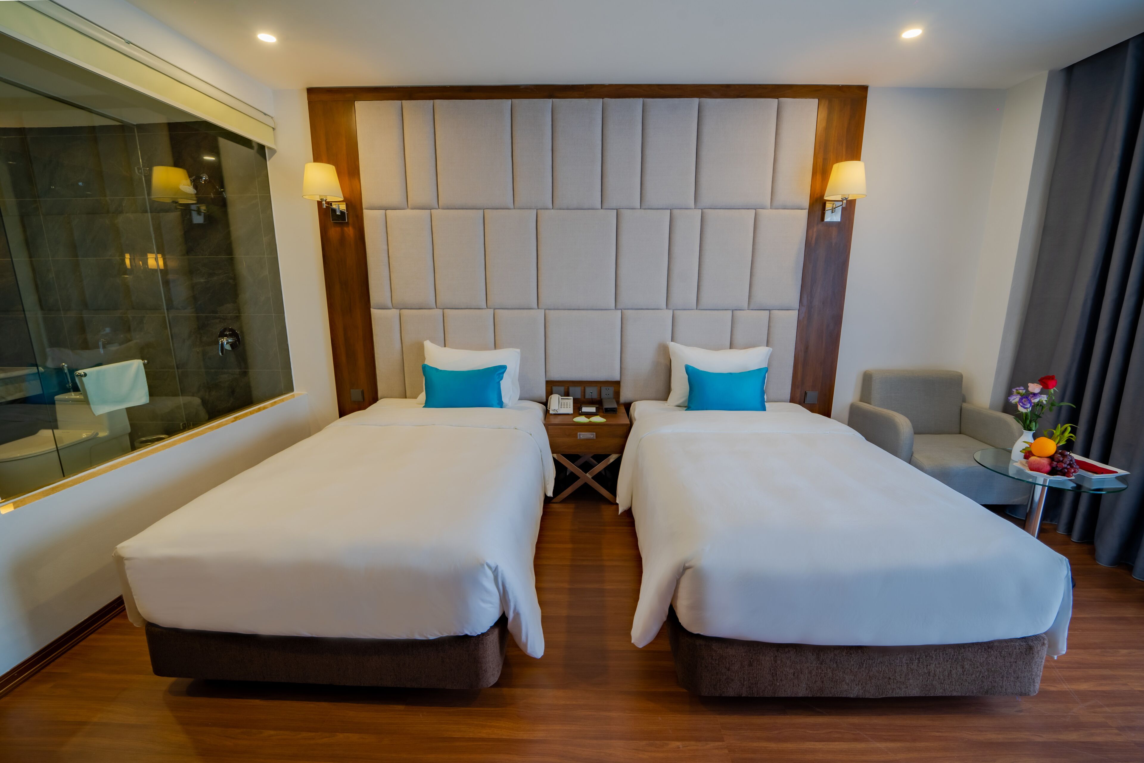 Een hotelkamer met twee bedden, een houten hoofdbord, een klein tafeltje met een telefoon en een lamp, en een glazen scheidingswand.