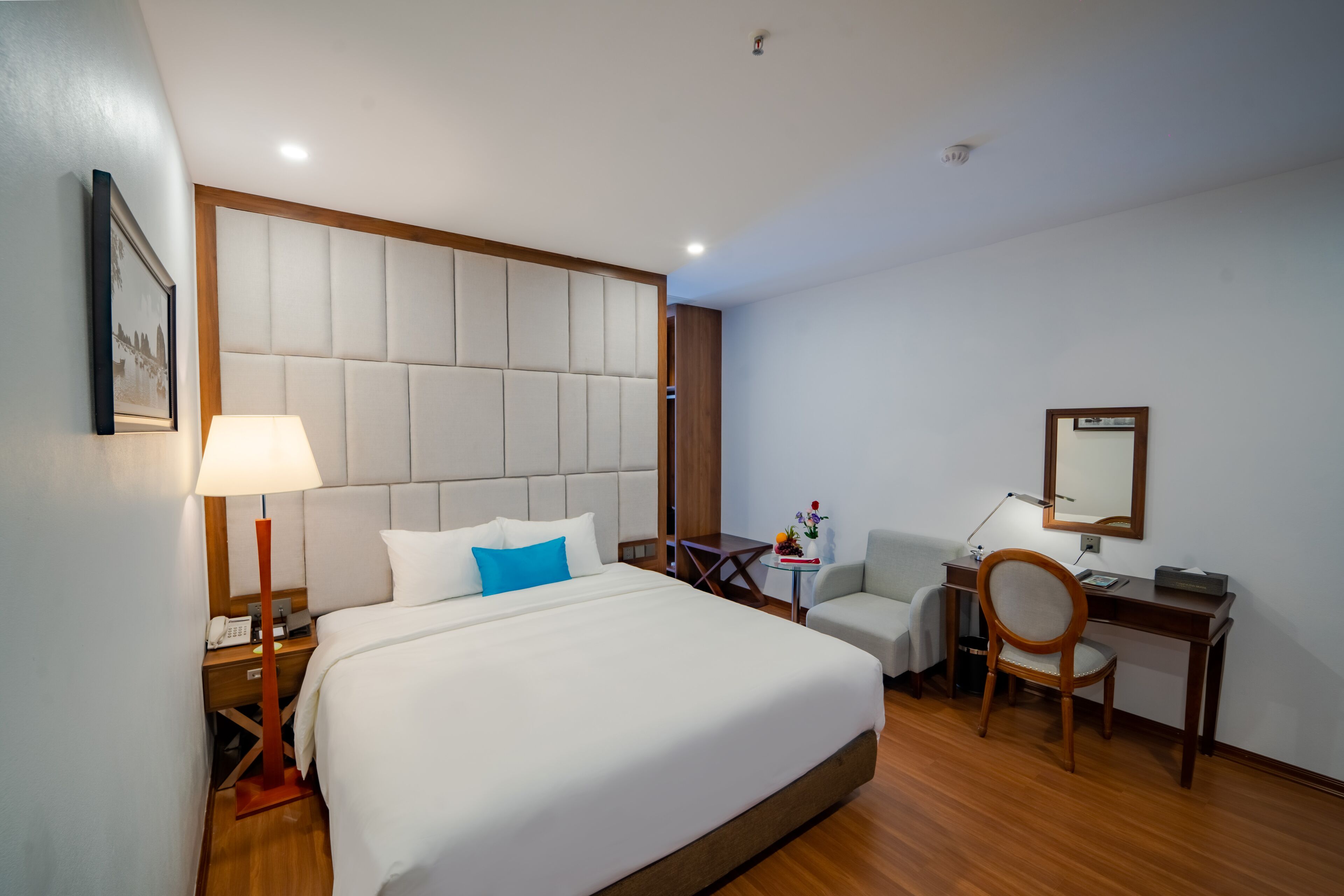 Habitación de hotel con una cama grande, un escritorio, una silla, una lámpara y una mesita con un jarrón de flores.