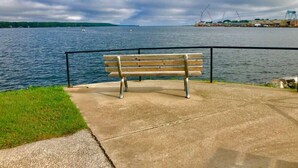 A concrete bench by a waterfront with a metal railing, overlooking a body of water and industrial structures in the distance. - Beach, harbor & restaurants within walking distance. Family friendly home! (Sturgeon Bay)
