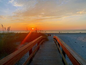 Een houten looppad dat naar een strand leidt, met een zonsondergang op de achtergrond.