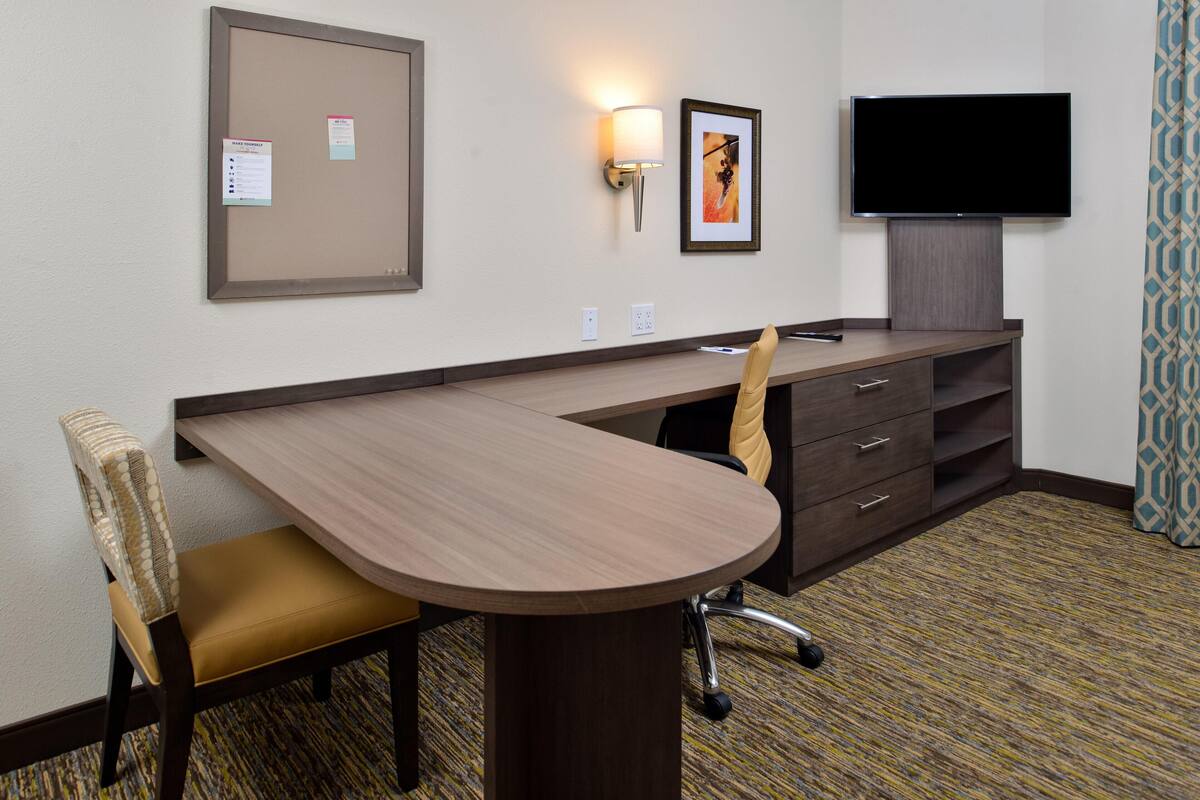 A hotel room with a desk, chair, television, and a bulletin board.