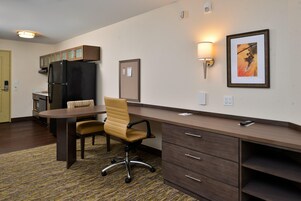 Una habitación de hotel compacta con kitchenette, un comedor con mesa y sillas, un escritorio con cajones y un cuadro en la pared.