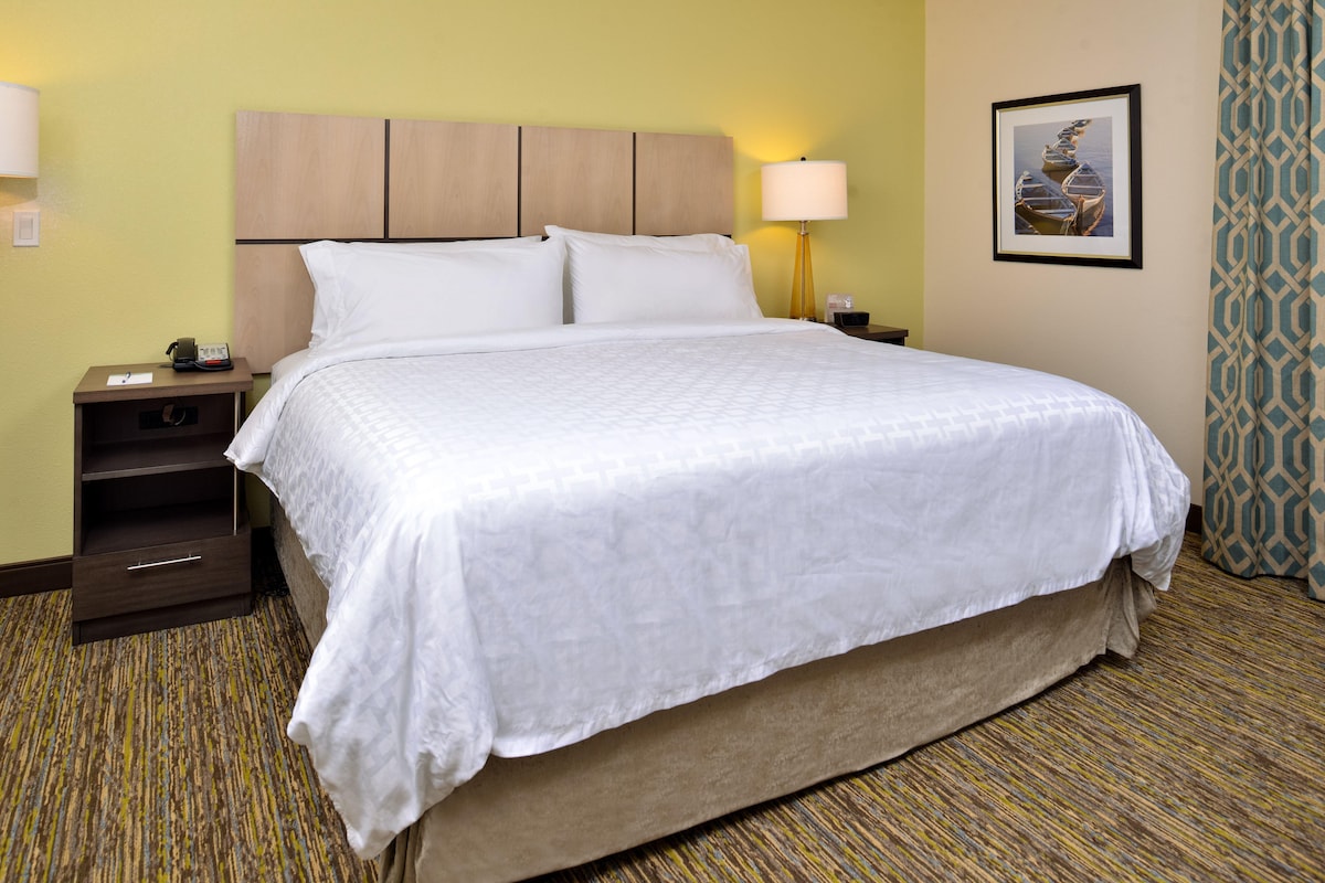 A hotel room with a large bed, a nightstand with a lamp, a framed picture on the wall, and a patterned carpet.