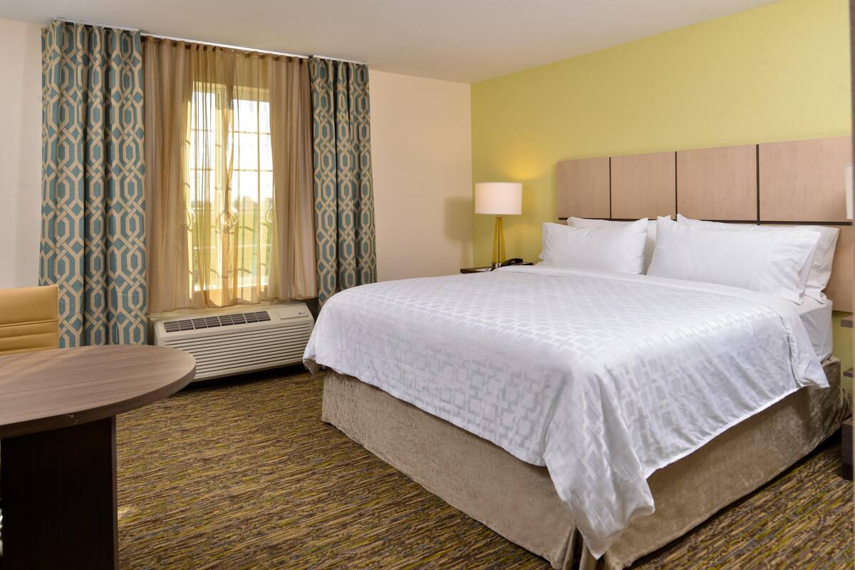A hotel room with a large bed, a bedside table, a lamp, and a window with curtains.