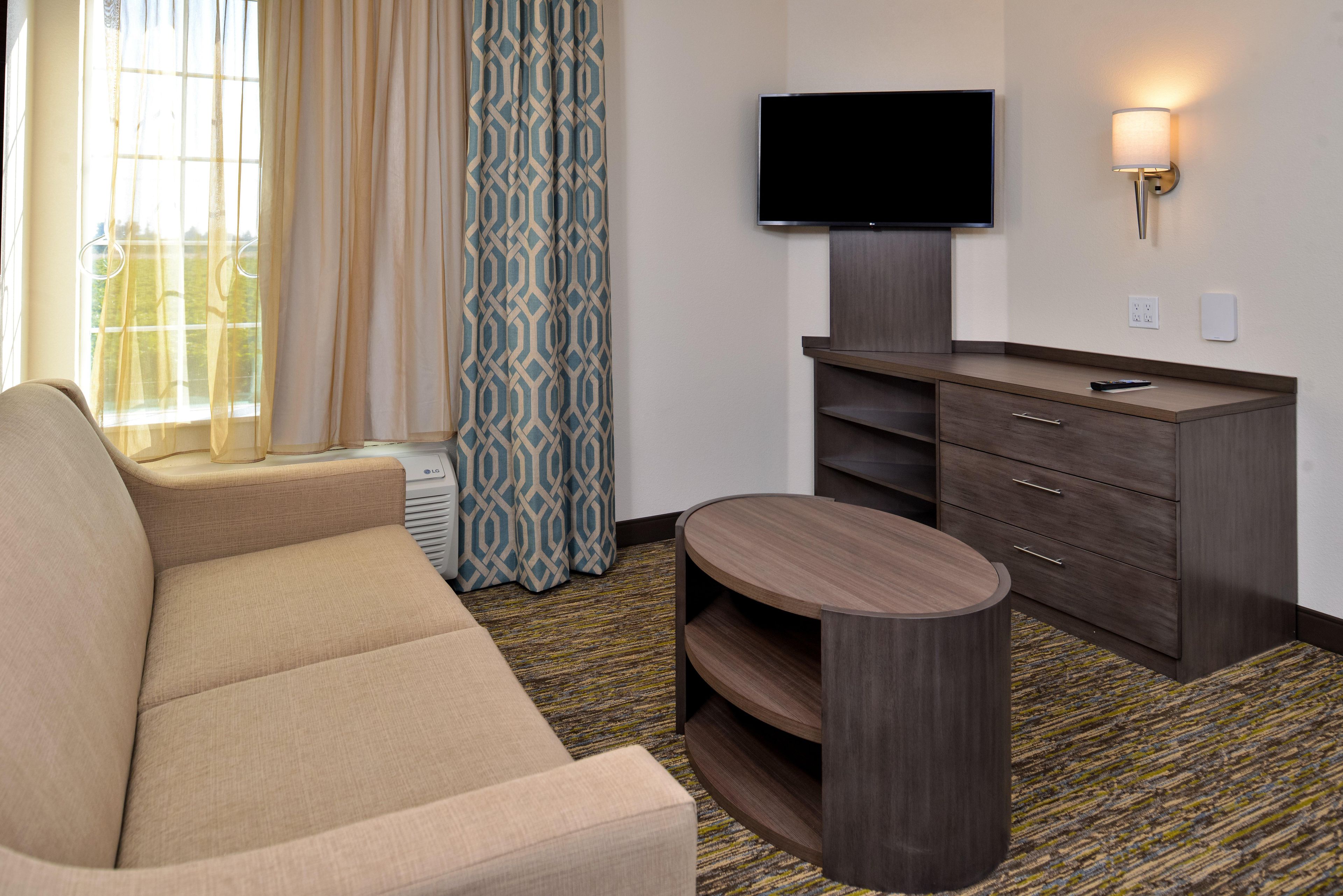 A hotel room with a sofa, a TV mounted on the wall, a desk with drawers, and a patterned carpet.