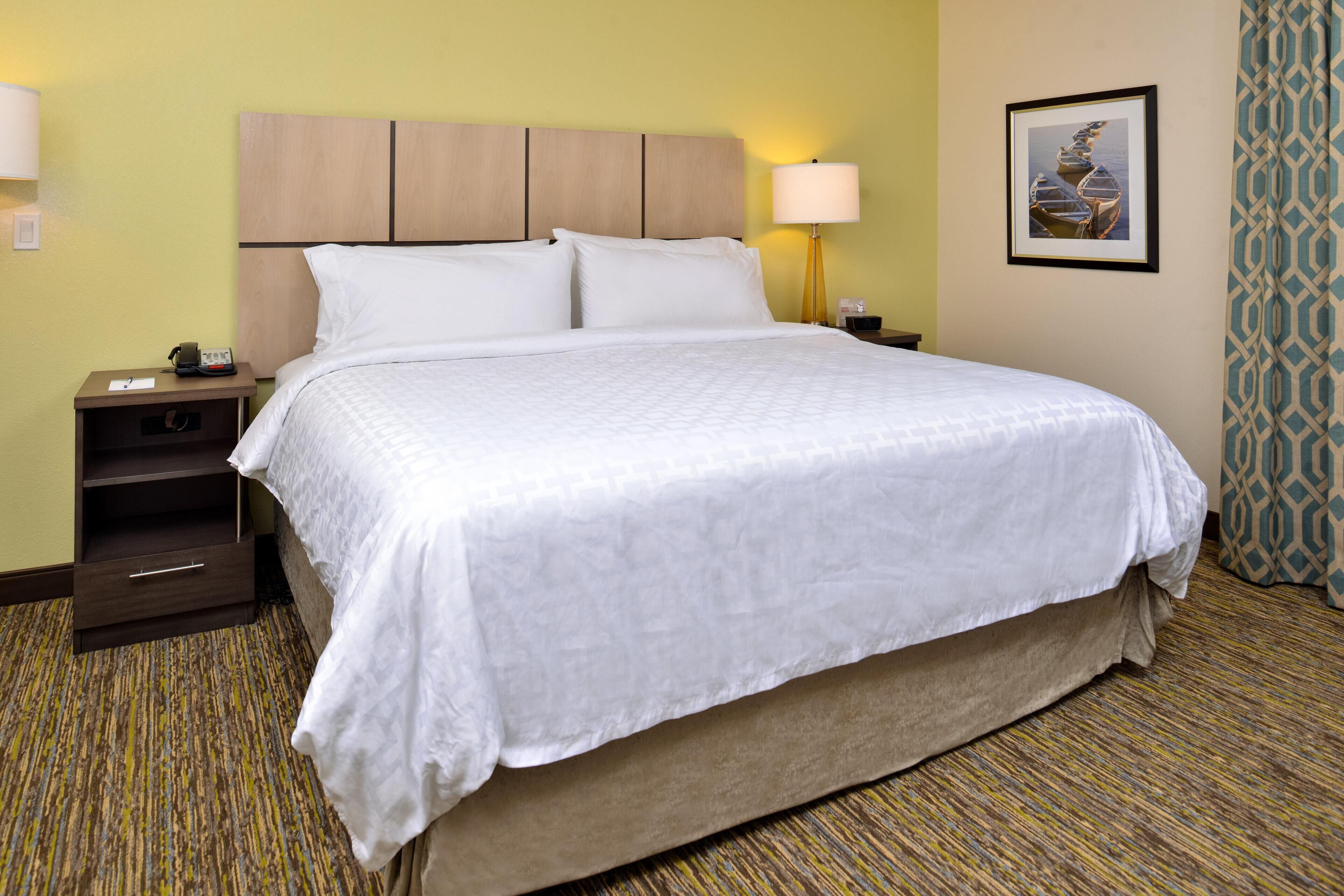 A hotel room with a large bed, a nightstand with a lamp, a framed picture on the wall, and a patterned carpet.