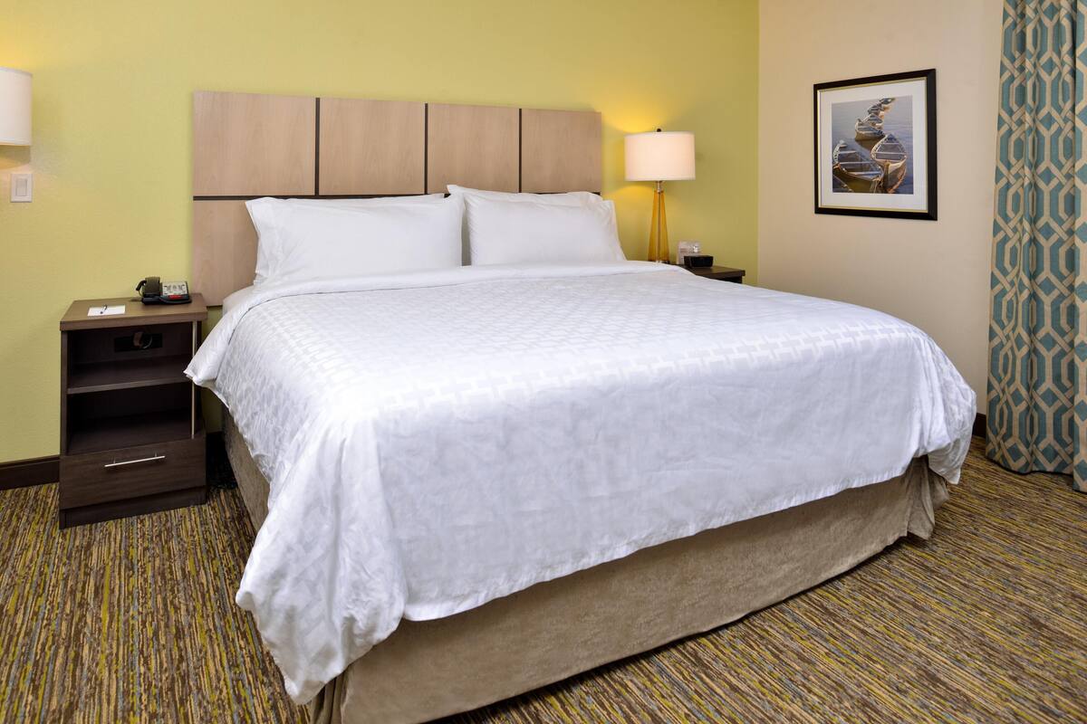 A hotel room with a large bed, a nightstand with a lamp, a framed picture on the wall, and a patterned carpet.