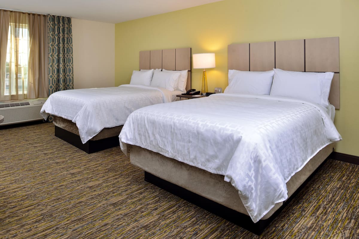 A hotel room with two beds, a bedside table with a lamp, and a window with curtains.