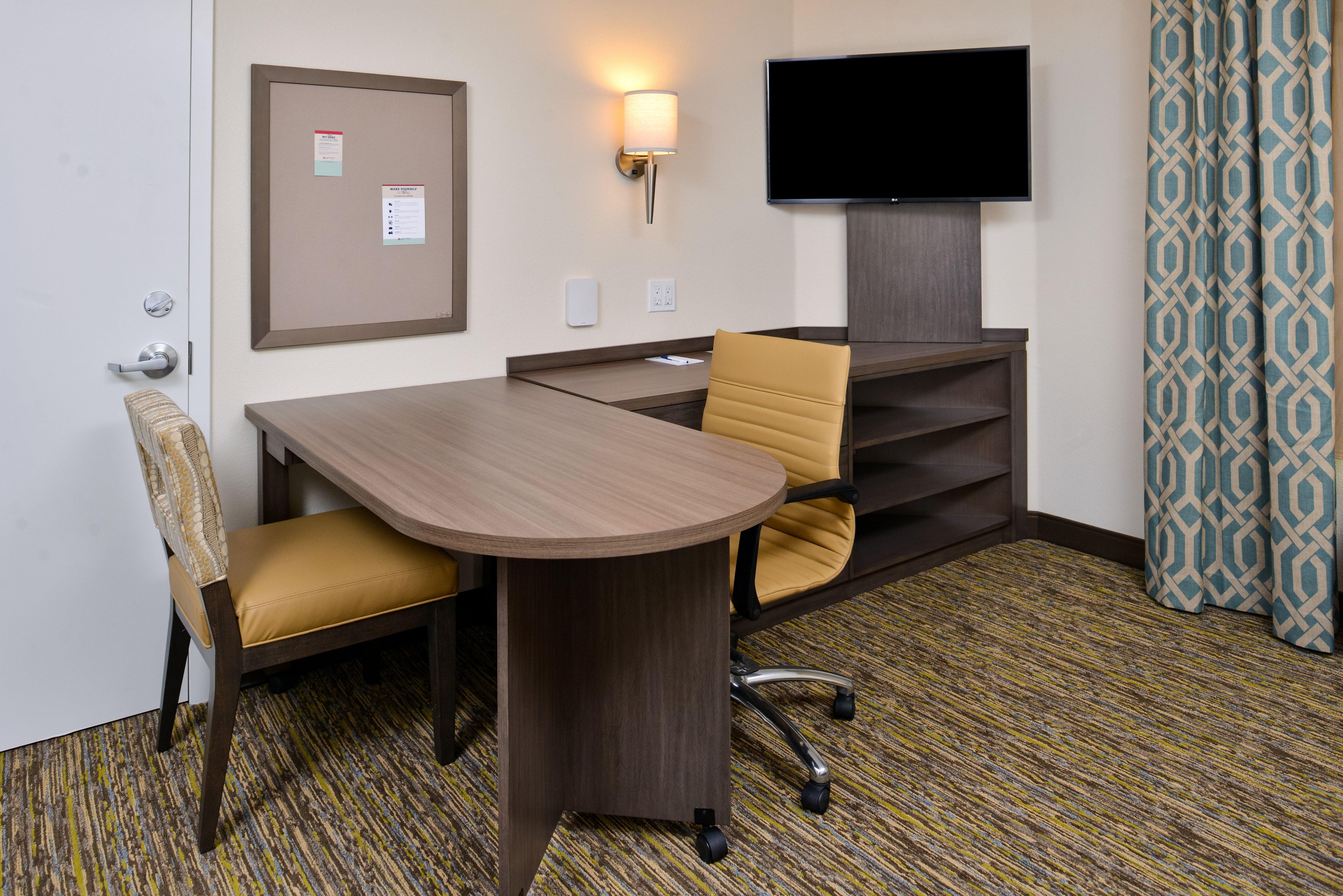 A hotel room with a wooden desk, two chairs, a television, a bulletin board, and patterned curtains.