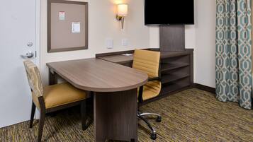 A hotel room with a wooden desk, two chairs, a television, a bulletin board, and patterned curtains.