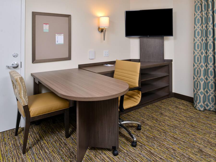 A hotel room with a wooden desk, two chairs, a television, a bulletin board, and patterned curtains.