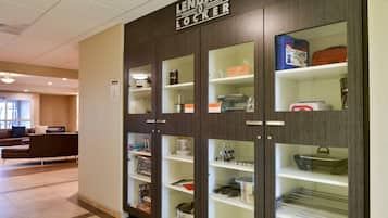Una hall d'albergo moderna con un'area deposito che include porte in vetro, scaffali con utensili da cucina e un angolo relax con divano e tavolino.