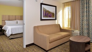 A hotel room with a bed, a sofa, a round wooden side table, a framed picture on the wall, and a patterned curtain. - Candlewood Suites Lodi (Lodi)